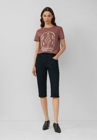 Braunes Grafik-T-Shirt mit weißem Druck und kurzen Ärmeln, kombiniert mit dunklen, hochgekrempelten Chinos. Das Modell trägt schwarze Slipper.