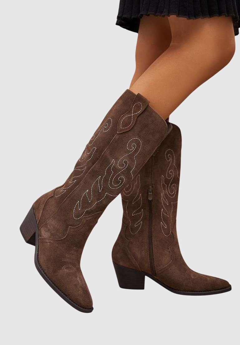 Bottes en daim marron jusqu'aux genoux avec des détails brodés et des talons carrés, portées avec une jupe noire sur un fond uni.