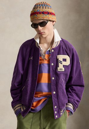 Jeune homme portant un bonnet en tricot coloré, des lunettes de soleil foncées, une veste universitaire violette, une chemise rayée orange et violette, un pantalon vert et un collier de perles.