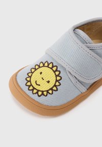Chaussure en tissu bleu clair avec une semelle en caoutchouc tan, arborant un motif de tournesol brodé jaune sur le bout et une sangle en Velcro.
