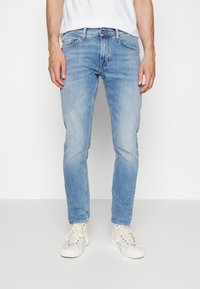 Jeans azul claro combinados con una camiseta blanca y zapatillas crema, de pie contra un fondo blanco liso. Manos relajadas a los lados.