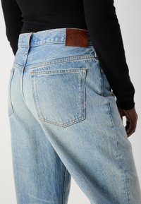 Jeans en denim bleu clair avec une taille haute, deux poches arrière et un patch en cuir marron de la marque. Texture légèrement distendue au niveau des coutures.