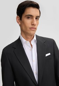 Giacca nera con una camicia bianca sotto. La camicia ha un colletto a punta e dettagli con bottoni, con un fazzoletto nella tasca sinistra.