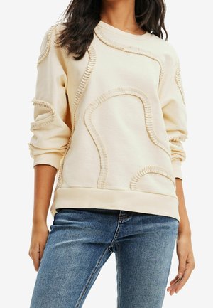 Kobieta w kremowej bluzie z abstrakcyjnymi, przeszywanymi wzorami i niebieskich dżinsach, z ramionami swobodnie opuszczonymi wzdłuż ciała.