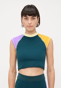 Kvinne med kort mørkt hår, som har på seg hoop-øreringer og en ermeløs fargeblokket crop top i turkis, lilla, gul og lys gul.