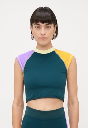 Femme avec les cheveux courts et foncés portant des boucles d'oreilles créoles et un crop top sans manches en coloris bloc vert turquoise, violet, jaune et jaune clair.