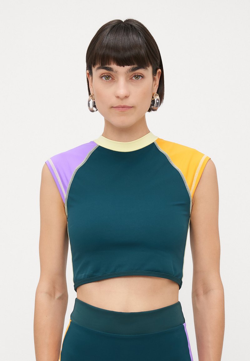 Kvinne med kort mørkt hår, som har på seg hoop-øreringer og en ermeløs fargeblokket crop top i turkis, lilla, gul og lys gul.