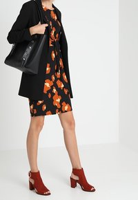 Blazer noir sur une robe noire à motif floral orange, accompagné d'un sac en cuir noir et de bottines à talons rouges.