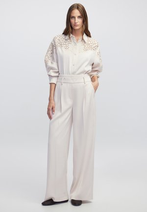 Camicia button-up color beige chiaro con dettagli in pizzo sulle spalle, abbinata a pantaloni a gamba larga dello stesso colore e dalla texture morbida e liscia.