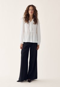 Woman with curly hair wearing a white long-sleeve blouse and wide-leg black pants standing against a neutral background.