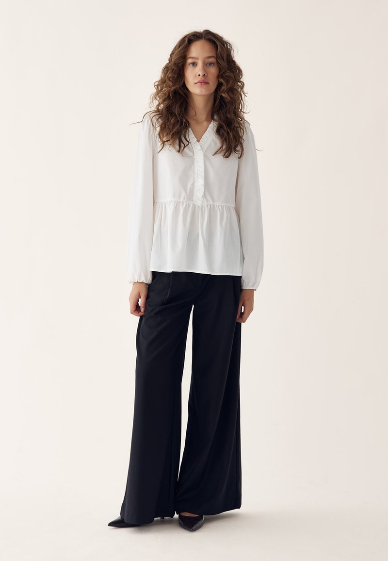 Woman with curly hair wearing a white long-sleeve blouse and wide-leg black pants standing against a neutral background.