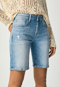 Shorts de mezclilla azul claro con un corte ajustado, ligeras desgastaciones y dobladillo enrollado. Combinados con un top de punto beige, exhibiendo un estilo casual.