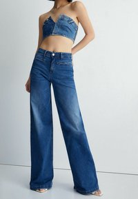Kvinna som bär högmidjade vida blå jeans och en strapless denim crop top med dragkedja och knappdetaljer, stående på vit golv.