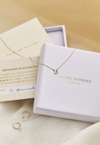 Collier en or avec un pendentif en forme de lettre "B" sur une chaîne, accompagné de boucles d'oreilles créoles ornées de perles et d'un certificat dans un emballage blanc.