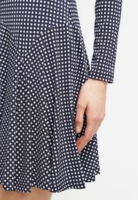 Marinblå klänning med vitt rutmönster; figursydda långärmade ärmar och veckad kjoldel; mjukt, texturerat tyg; hand vilande på kjolen.