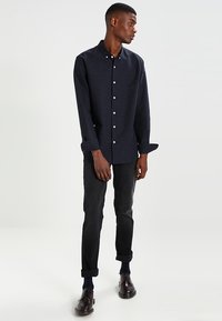 Camicia button-up blu scuro con un motivo a macchie, maniche lunghe e colletto. Indossata con jeans scuri arrotolati e scarpe marroni.