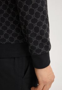 Czarny sweter z szarym geometrycznym wzorem, z prążkowanym mankietem i dołem. Tkanina wydaje się strukturalna, podkreślająca skomplikowany wzór.