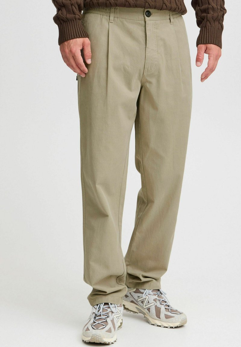 Beige trousers with a relaxed fit, featuring pleats, belt loops, and side pockets. Worn with light gray and brown patterned sneakers.