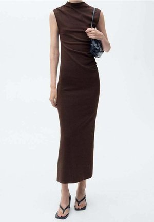 Kobieta w ciemnobrązowej, dopasowanej sukience na ramiączkach o długości midi, trzymająca małą czarną torebkę na ramię i nosząca czarne sandały na obcasie.