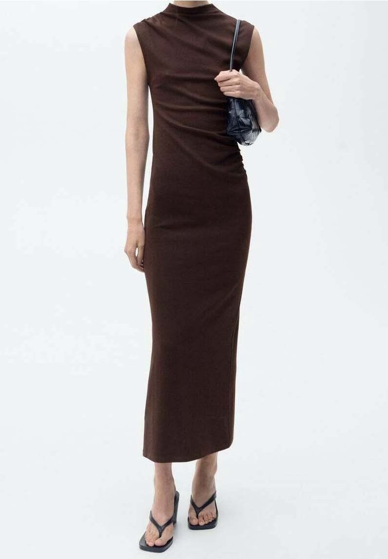 Femme portant une robe midi sans manches, moulante, de couleur brun foncé, tenant un petit sac à épaule noir et portant des sandales à talons noires.