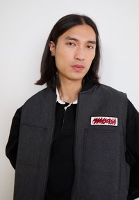 Gray vest with a zippered front, two front pockets, and a patch. Layered over a black long-sleeve shirt with a white collar.