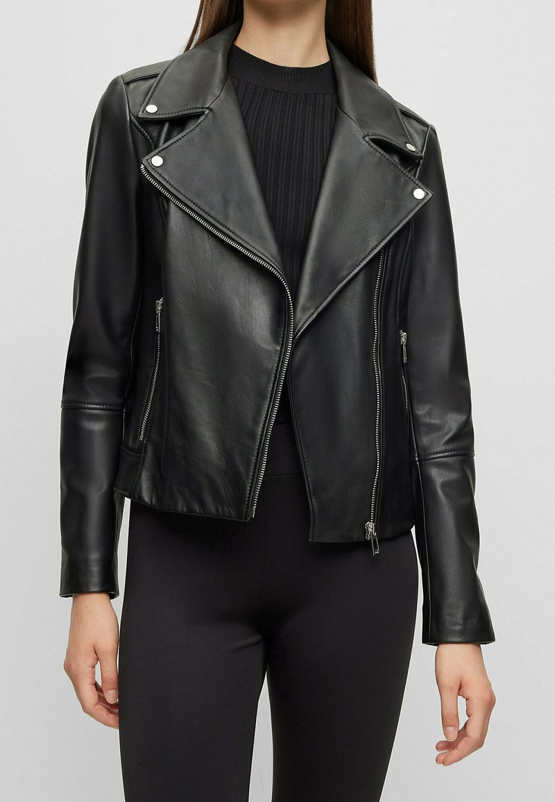 Mujer con una chaqueta de cuero negra ajustada de estilo biker sobre una camiseta negra de canalé, combinada con pantalones negros, de pie frente a un fondo claro y sencillo.