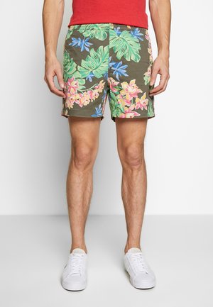 Mand iført shorts med blomsterprint i grønne, blå, pink og gule blade og blomster, hvide sneakers og en rød skjorte, stående på hvidt gulv.