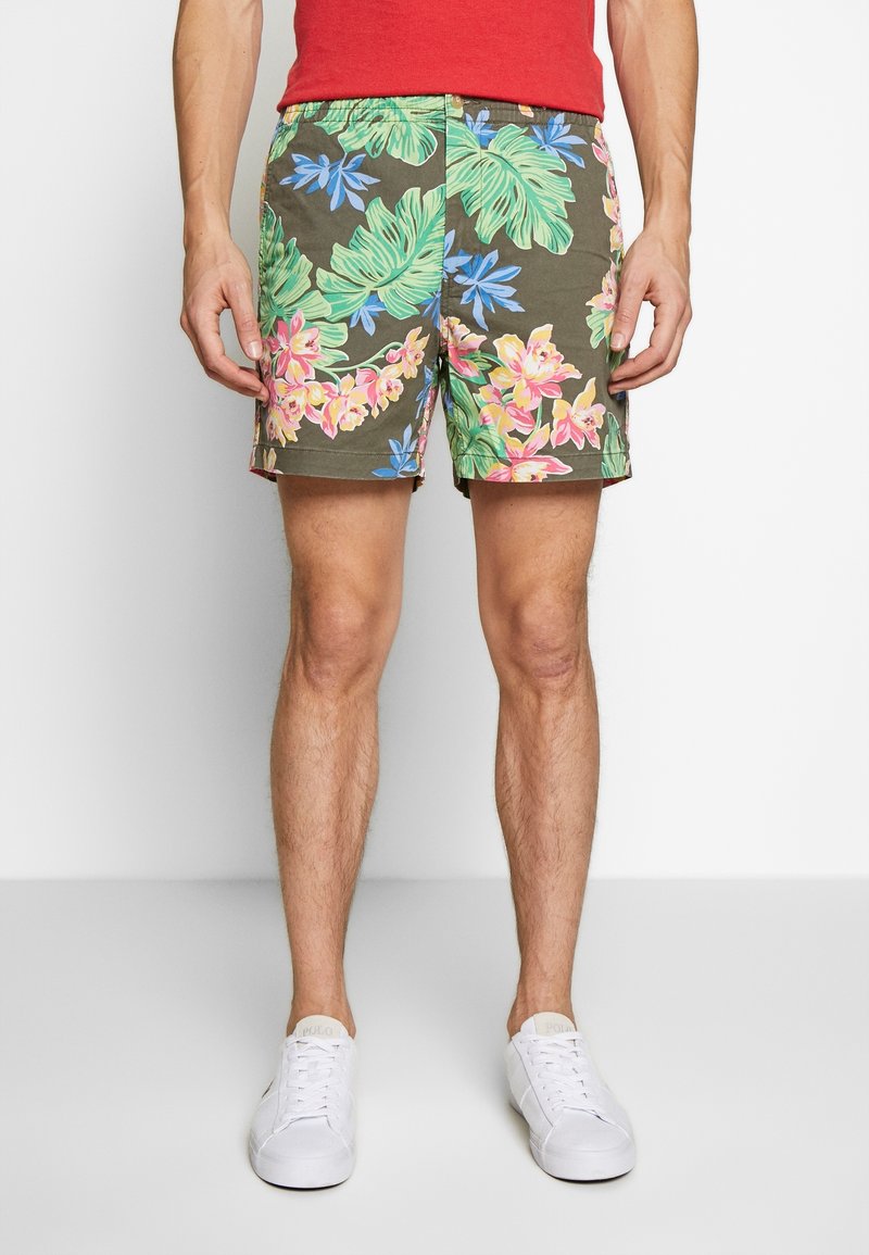 Homme portant un short à imprimé floral avec des feuilles et des fleurs vertes, bleues, roses et jaunes, des baskets blanches et une chemise rouge, debout sur un sol blanc.