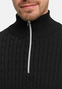 Musta neulepaita, jossa on korkea resorikaulus, vetoketjukiinnitys edessä ja teksturoitu palmikkoneulekuvio. Sileä viimeistely ja istuva leikkaus.