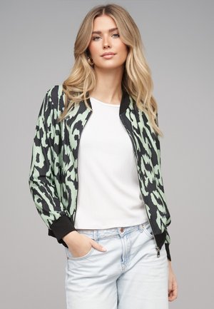 Donna bionda che indossa una giacca bomber verde e nera con stampa animalier, maglietta bianca, jeans azzurri, in piedi con una mano in tasca.