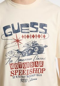 T-shirt in cotone color crema con stampa grafica audace in blu e rosso, raffigurante un'auto d'epoca e elementi testuali, scollatura rotonda, maniche corte.