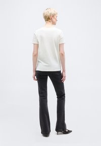 Άτομο με κοντά ξανθά μαλλιά, που φαίνεται από πίσω, φοράει λευκό κοντομάνικο πουκάμισο, μαύρο καμπάνα τζιν και μαύρα παπούτσια με τακούνι.