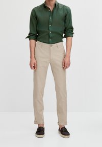 Mężczyzna w ciemnozielonej koszuli na guziki z podwiniętymi rękawami, beżowych spodniach z podwiniętymi nogawkami i czarnych espadrylach na slip-on, stojący na jednolitym tle.