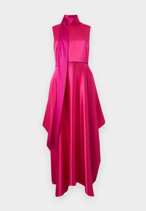 Femme debout dans une robe en satin sans manches rose vif avec détail d'écharpe à col montant et chaussures marron à talons sur fond blanc.
