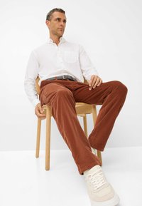 Chemise blanche à boutons avec une poche, pantalon en velours côtelé marron, ceinture grise et baskets crème. Posture assise sur une chaise en bois devant un fond uni.