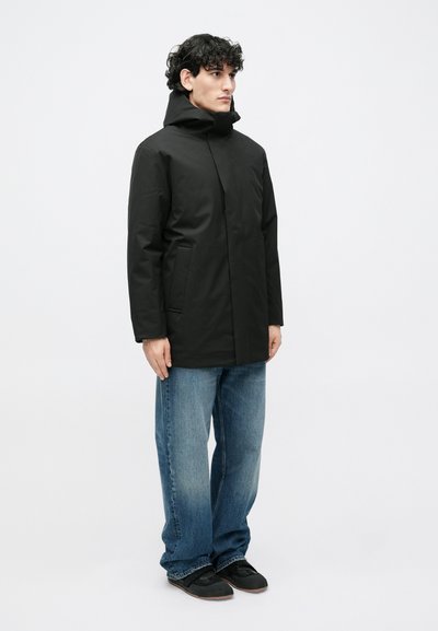 Jachetă impermeabilă neagră cu guler înalt, buzunare frontale și glugă; combinată cu blugi largi albaștri și adidași negri.