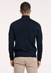 Maglione blu scuro con maniche lunghe, colletto e orlo a coste. Presenta una texture a maglia liscia. Indossato con pantaloni beige.