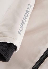 Šviesiai smėlio spalvos striukė su juodu raštuotu rankogaliu, išraiškingu pilku "SUPERDRY®" logotipu ir juodu užtrauktuko detalė.