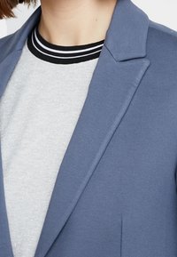 Niebieska marynarka o zbudowanej konstrukcji, z klapami i gładkim materiałem. Noszona na jasnoszarym koszuli z czarno-białym paskiem przy kołnierzyku.