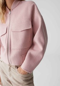 Veste zippée rose en matière douce avec deux poches avant, col et poignets côtelés. Forme arrondie avec épaules tombantes.
