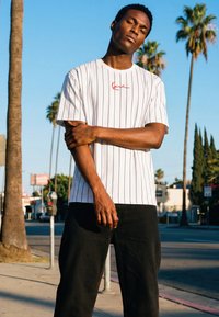 Ung mand står på en solrig gade med palmer, iført en hvid stribet T-shirt og sorte bukser, med armene afslappet krydset.