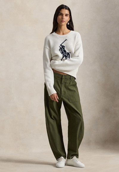 Άσπρο πλεκτό πουλόβερ με μπλε λογότυπο αλόγου, σε συνδυασμό με άνετο πράσινο παντελόνι και άσπρα αθλητικά παπούτσια. Μαλακή υφή και χαλαρή εφαρμογή.