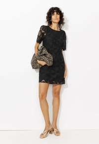 Robe en dentelle noire à manches courtes, motif floral, coupe ajustée. La mannequin tient un sac en imprimé léopard. Portée avec des talons nus à lanières.