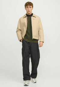 Beige bomber jacket, khaki green sweater, black cargo pants, and silver sneakers. Sleek design with a zippered chest pocket. Textured materials.
