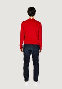 Maglione rosso con polsini a costine, abbinato a jeans blu scuri. L'outfit è completato da sneakers bianche. Etichetta del marchio visibile sul maglione.