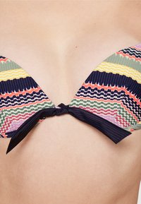 Top de biquíni multicolorido com um padrão ondulado em verde, amarelo, azul-marinho e rosa. O tecido é canelado, com um fecho de laço preto nas costas.