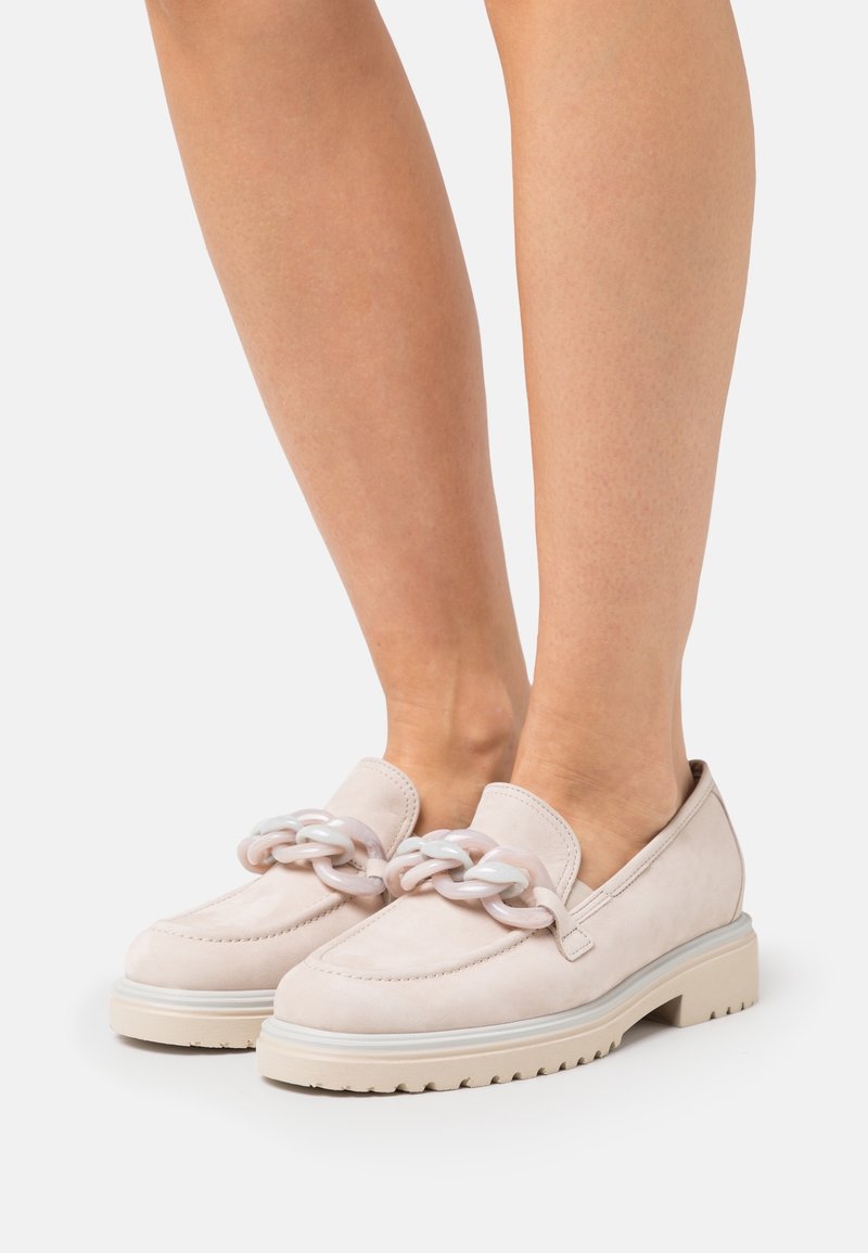 Pieds portant des mocassins beige clair avec un grand détail brillant de chaîne sur le dessus et une semelle épaisse, présentés sur un fond uni.