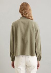 Olijfgroene lichtgewicht blouse met een kraag en gebloemde lange mouwen, met een losse pasvorm die net boven de taille eindigt. Witte jeans zichtbaar.