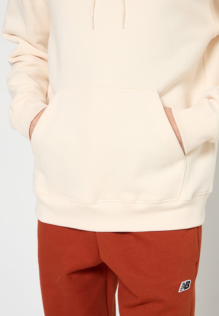 Hoodie couleur crème en tissu doux avec une poche kangourou, une capuche avec cordon de serrage, des manches à revers, associé à un pantalon couleur rouille arborant un logo.