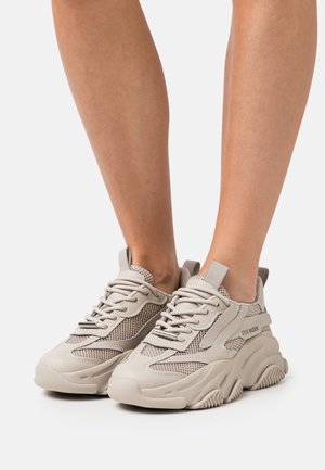 Light beige sneakers featuring a mix of smooth and mesh textures, thick soles, and tonal laces, with a prominent brand logo on the side.